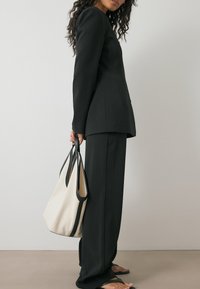 Mustat räätälöidyt puvut, joissa on pitkät hihat ja suorat housut, yhdistettynä beigelle olkalaukulle, jossa on mustia yksityiskohtia, neutraalia taustaa vasten.