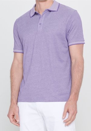 Uomo che indossa una polo lilla a maniche corte con colletto e polsini a righe, abbinata a pantaloni bianchi, in piedi davanti a uno sfondo uniforme.