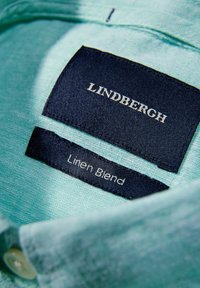 Jasno miętowa koszula z lnu z kołnierzykiem, z granatową metką wyświetlającą napis "LINDBERGH" i "Linen Blend" w białym tekście.