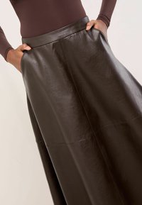 Barna műbőrből készült, magas derekú szoknya, sima textúrával, széles fazonnal és oldalsó zsebekkel. Nincsenek látható záródások.