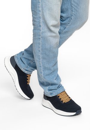 Person trägt hellblaue Jeans und schwarze Stricksneaker mit beige-farbenen Schnürsenkeln und geht auf weißem Hintergrund.