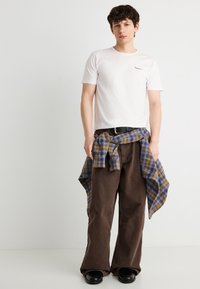 Νεαρός άνδρας που φοράει λευκό εφαρμοστό μπλουζάκι, καφέ παντελόνι με φαρδιά μπατζάκια, μαύρα παπούτσια, με μπλε και μουσταρδί καρό πουκάμισο δεμένο γύρω από τη μέση.