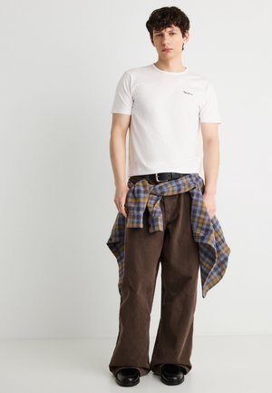 Mladý muž oblečený v bílém přiléhavém tričku, hnědých kalhotách s širokými nohavicemi, černých botách a s modro-hořčicovou kostkovanou košilí uvázanou kolem pasu.