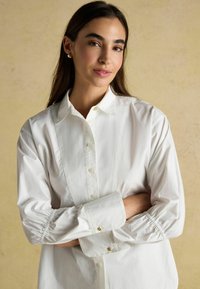 Weißes Hemd mit Knöpfen und lockerer Passform, mit elastischen Ärmeln und goldenen Knopfakzenten. Glatte Textur, klassisches Kragen-Design.