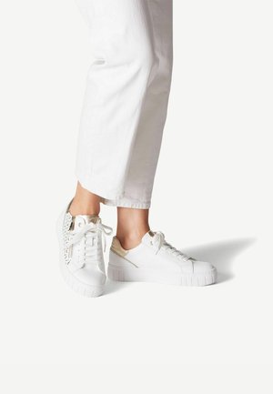 Vita sneakers med texturerad ytbeläggning, guldaccenter och platt gummisula. Snören och sömmar synliga; i kombination med vita korta byxor.