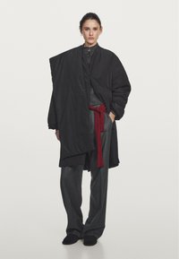 Cappotto nero oversized in tessuto texturizzato, design drappeggiato e tasche laterali; abbinato a pantaloni grigi a gamba larga e una cintura a strisce rosse.