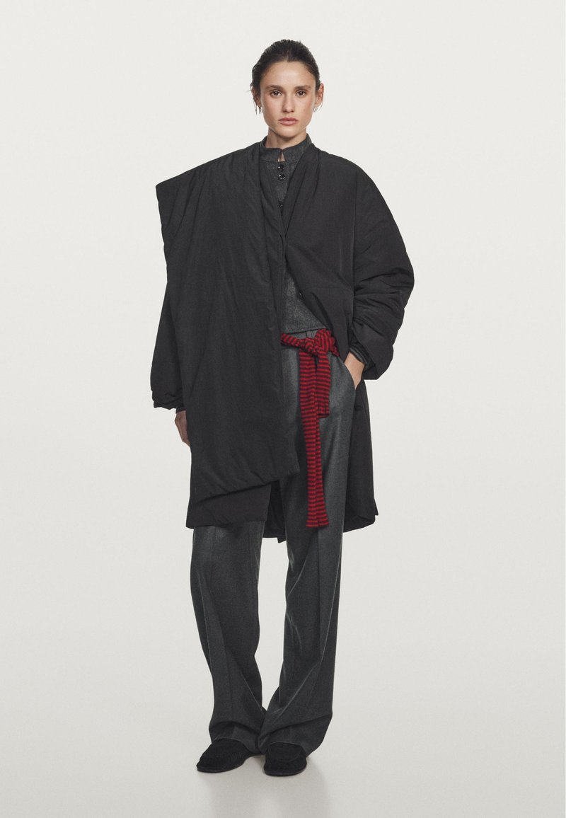 Cappotto nero oversized in tessuto texturizzato, design drappeggiato e tasche laterali; abbinato a pantaloni grigi a gamba larga e una cintura a strisce rosse.