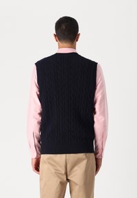 Σκούρο μπλε αμάνικο πουλόβερ με πλέξη κασκόλ, με ριγωτό στρίφωμα, φορεμένο πάνω από ένα ανοιχτό ροζ πουκάμισο με κουμπιά, διαθέτοντας κλασικό σχέδιο.