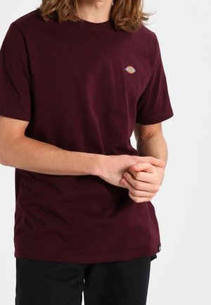 Personne aux cheveux longs portant un t-shirt bordeaux foncé à manches courtes et un pantalon noir, les mains jointes, debout devant un fond uni.