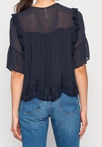 Blusa trasparente blu navy con maniche a volant, design plissettato e ricami floreali lungo l'orlo. Indossata sopra jeans in denim.