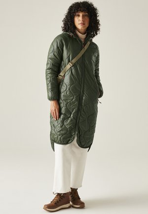 Mujer con cabello rizado que lleva una chaqueta larga acolchada verde, pantalones blancos, botas de senderismo marrones y un bolso cruzado, de pie contra un fondo liso.