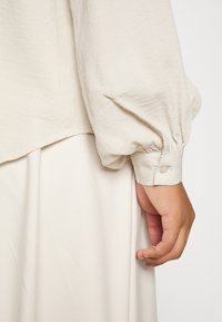 Ljus beige blus med puffärmar och knäppta manschetter, i kombination med en krämfärgad kjol med mjukt, texturerat tyg.