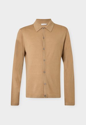 Man met krullend haar die een beige gebreid vest met knopen draagt, een wit overhemd, losse blauwe spijkerbroek en witte sneakers, staat met handen in de zakken.