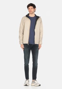 Giacca leggera beige con cappuccio nero, zip dorata, indossata sopra una maglietta blu navy. Abbinata a jeans scuri e sneakers grigie.