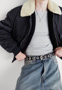 Chaqueta bomber negra con un cuello de borrego beige, camisa gris ajustada, pantalones de mezclilla azules y un cinturón negro con remaches y hebilla plateados.