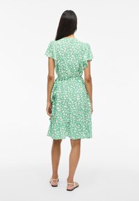 Grønn kjole med korte flakkende ermer, prydet med et penselstrøk mønster i kremfarge. Strukturert stoff med A-linjeform og volangkant.