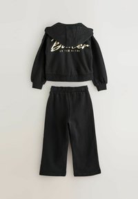 Sudadera negra con capucha y cinturilla elástica, que presenta "Baker" en dorado metálico en la parte trasera. Combinada con pantalones de chándal negros a juego.