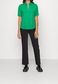 Femme portant une chemise à boutons à manches courtes verte, un pantalon noir droit et des chaussures à lacets noires à plateforme, se tenant contre un fond blanc.