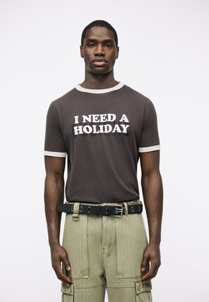 Άνδρας που φοράει μαύρο μπλουζάκι με λευκές λεπτομέρειες και κείμενο «ΧΡΕΙΑΖΟΜΑΙ ΔΙΑΚΟΠΕΣ», συνδυασμένο με πράσινο παντελόνι cargo και μαύρη ζώνη.