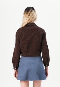 Kvinne med langt blondt hår iført en brun leopardmønstret jakke og et blått denimskjørt, sett bakfra mot en ensfarget bakgrunn.