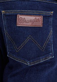 Ciemnoniebieska dżinsowa kieszeń z brązową skórzaną etykietą "Wrangler". Posiada żółte szwy oraz charakterystyczny zygzakowaty wzór na kieszeni.