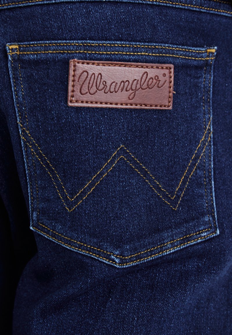 Tasca in denim blu scuro con etichetta in pelle marrone "Wrangler". Presenta cuciture gialle e un distintivo motivo a zig zag sulla tasca.