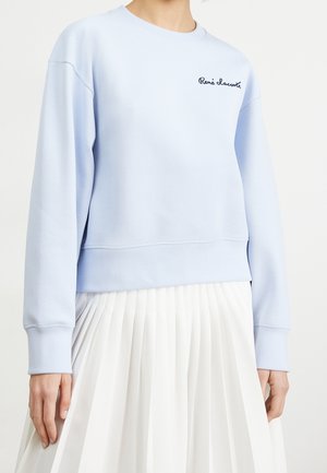 Kobieta nosząca jasnoniebieską bluzę z długim rękawem z haftem „René Lacoste”, połączoną z białą plisowaną spódnicą z wysokim stanem.