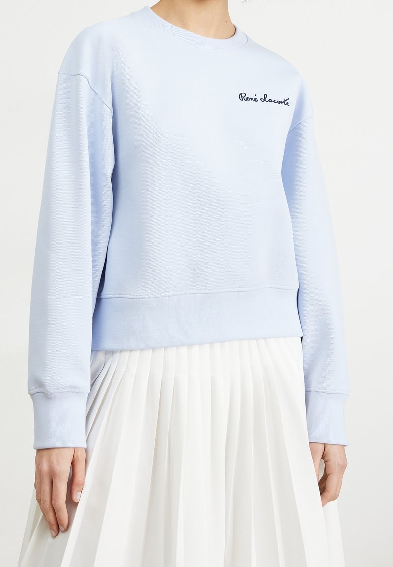 Femme portant un sweat-shirt bleu clair à manches longues avec une broderie « René Lacoste », associé à une jupe plissée blanche taille haute.