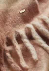 Textil din amestec de lână cu nuanțe moi de roz și crem, având o textură pufoasă și un mic etichetă metalică cu un logo încorporat în țesătură.