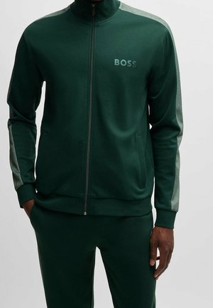 Dunkelgrüne Trainingsjacke mit hellgrauen Streifen an den Ärmeln, Frontreißverschluss, Seitentaschen und „BOSS“-Logo auf der linken Brust, getragen mit passenden Hosen.