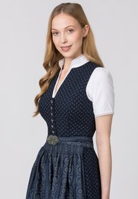 Traditionelles Dirndlkleid mit einem dunkelblauen gemusterten Mieder, einer weißen Bluse mit Spitzendetails und einem gerafften Rock mit einer dekorativen Gürtelschnalle.