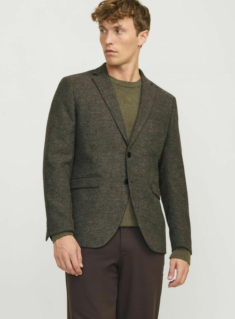 Blazer en tweed marron avec revers cranté, deux poches avant et un bouton. Porté par-dessus un pull vert olive et un pantalon marron.