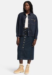 Chaqueta de mezclilla de botones, azul oscuro, con un bolsillo en el pecho. Combinada con una falda de mezclilla oscura con botones en la parte delantera y botas marrones con suelas gruesas.