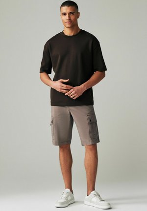 Tricou cu mânecă scurtă de culoare maro, pantaloni scurți cargo gri cu buzunare laterale și adidași albi. Textură netedă, croială lejeră, design casual.