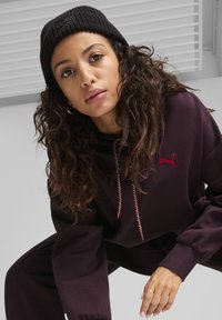 Mørk lilla hoodie med røde snore og et lille rødt katte-logo. Bæres med en sort strikket hue. Krøllet hår synligt, baggrunden er neutral.