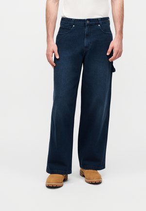 Mand iført mørkeblå vidde jeans, tan farvede støvler og en hvid indstukket skjorte, stående mod en ensfarvet lys baggrund.