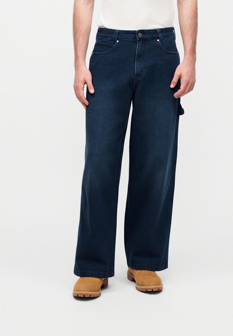 Homme portant un jean large bleu foncé, des bottes couleur camel et une chemise blanche rentrée, debout devant un fond clair uni.