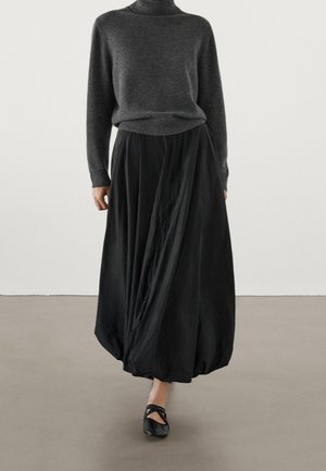 Femme portant un pull à col roulé gris foncé, une jupe boule noire et des ballerines noires marchant à l'intérieur sur un sol neutre.