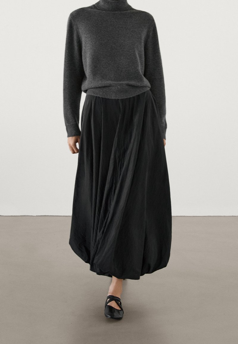Femme portant un pull à col roulé gris foncé, une jupe boule noire et des ballerines noires marchant à l'intérieur sur un sol neutre.