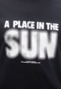 Μαύρο ύφασμα με λευκά κείμενα με τελείες που γράφουν "A PLACE IN THE SUN" και μικρότερο κείμενο από κάτω "MADS NØRGAARD - COPENHAGEN © 2026".