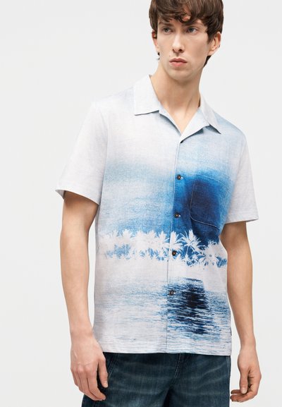 Νέος άνδρας που φοράει πουκάμισο με κοντά μανίκια με μπλε βαθμονόμηση και λευκό σχέδιο με φοίνικες και νερό, σε συνδυασμό με σκούρο τζιν.