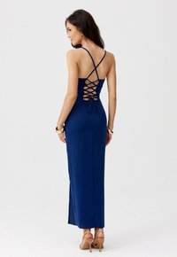 Žena v dlouhých námořnických šatech s křížovým šněrováním na zádech a tenkými ramínky, doplněná béžovými lodičkami na vysokém podpatku a zlatými náramky.