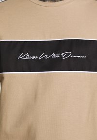 Béžové bavlněné tričko s vodorovným černým panelem a bílým vyšitým textem, který zní "Králové budou snít." Jednoduchý design s kontrastními barvami.