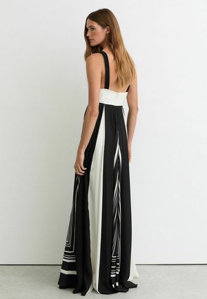 Moteris ilgais plaukais, dėvinti juodai baltą ilgaaulį suknelę be rankovių, stovinti šonu prie vientisos baltos sienos.