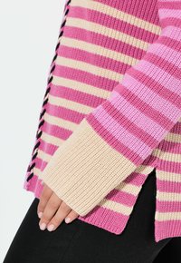 Stickad tröja med rosa och beige ränder, med ribbade ärmslut och sidoflikar, accentuerad med svarta snörningar längs sömmen.