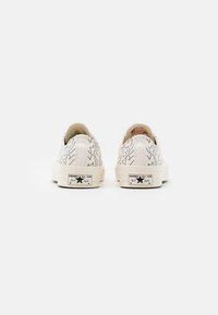 Bílé plátěné tenisky s černými abstraktními liniemi, gumovou špičkou a krémovými podrážkami; mají značku Converse All Star na patě.