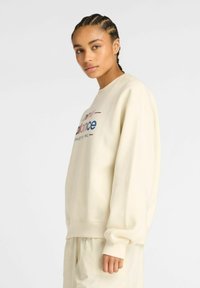 Jeune personne avec des cheveux tressés portant un sweat-shirt de couleur crème avec le logo "New Balance" et un pantalon crème assorti, se tenant légèrement de profil.