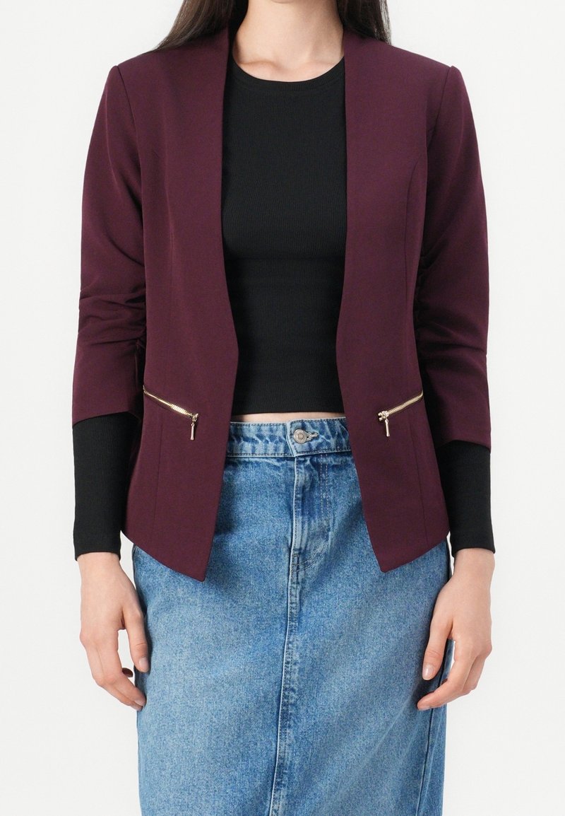 Femme portant un blazer violet foncé avec des fermetures éclair dorées sur un haut noir et une jupe en jean bleue sur un fond uni.