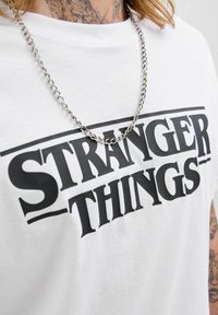Biała bawełniana koszulka z nadrukiem "STRANGER THINGS" wytłoczonym grubymi czarnymi literami, połączona ze srebrnym naszyjnikiem.