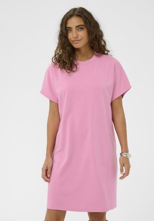 Moteris su garbanotais rudos spalvos plaukais, vilkinti laisvo stiliaus rožinę trumpų rankovių suknelę ir storą apyrankę ant dešinės riešo, stovinti priešais baltą foną.